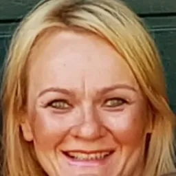 Portret uśmiechniętej blondynki, nauczycielki, z delikatnym makijażem i widocznymi zębami, na rozmytym tle. Ujęcie od przodu, skupione na twarzy.