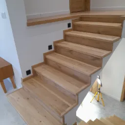 Nowoczesne, jasne schody drewniane z oświetleniem punktowym w ścianie, widoczne drzwi i lampa warsztatowa.