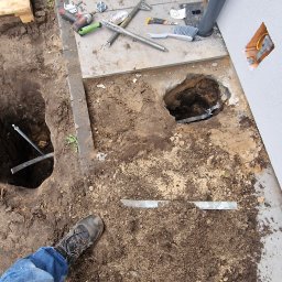 Canad Instalacje elektryczne - Wykopane doły pod instalację odgromową przy budynku. Widoczne narzędzia, fragment instalacji i kabel wystający ze ściany. Prace ziemne w toku.