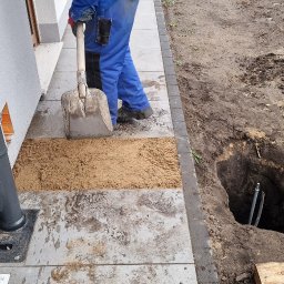 Canad Instalacje elektryczne - Prace ziemne przy budynku: wykop z rurami, łopata i piasek. Instalacja podziemna w trakcie realizacji, widoczne elementy instalacji elektrycznej.