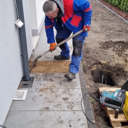 Canad Instalacje elektryczne - Pracownik w kombinezonie kopie dół pod instalację odgromową przy budynku. Widoczne narzędzia, skrzynka z akcesoriami i fragment żywopłotu.