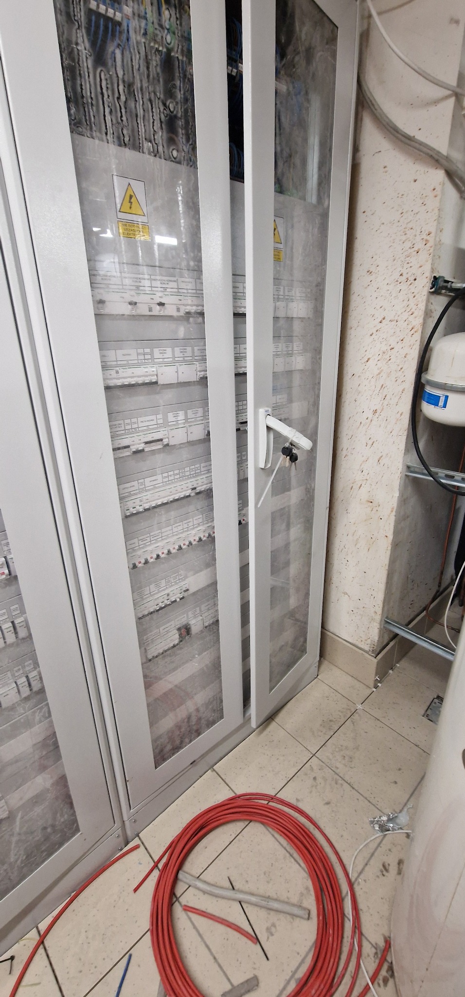 Szara szafa rozdzielcza z widocznymi zabezpieczeniami i okablowaniem wewnętrznym, kluczyki w zamku, zwinięty czerwony przewód na podłodze.