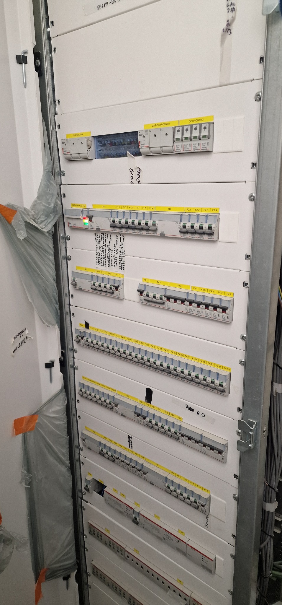 Rozdzielnica elektryczna z wieloma bezpiecznikami i wyłącznikami, widoczne oznaczenia faz i obwodów, częściowo zakryta folią ochronną, w tle widoczne kable.