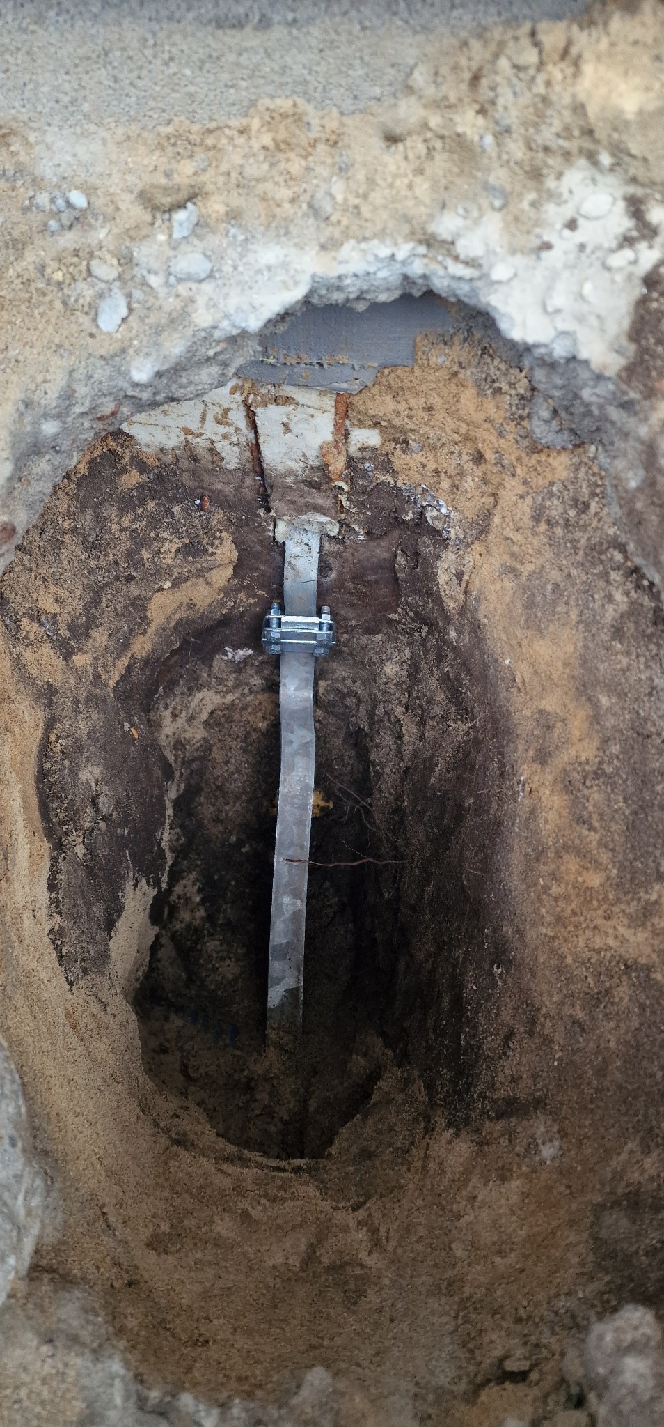 Uziemienie pionowe: stalowa szyna w wykopie, mocowana śrubami. Widoczne warstwy gruntu i fragment betonowej konstrukcji. Instalacja odgromowa w trakcie prac.
