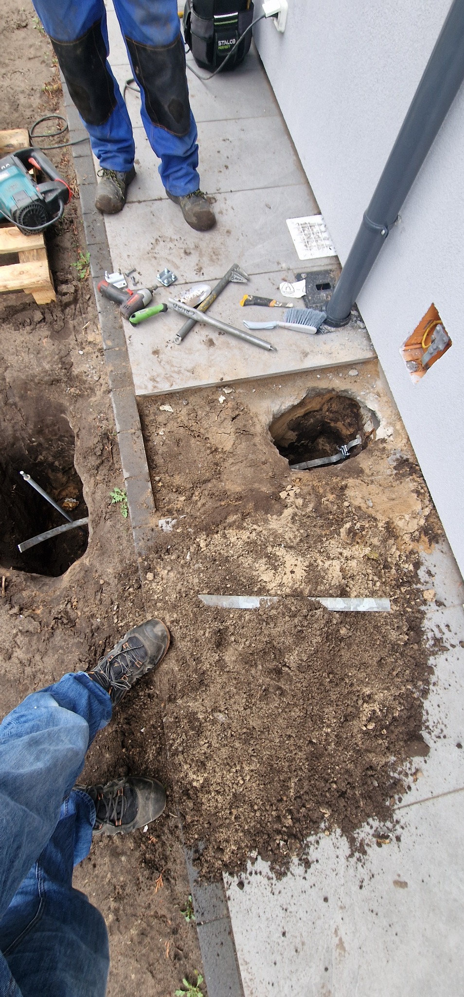 Wykopane doły pod instalację odgromową przy budynku. Widoczne narzędzia, fragment instalacji i kabel wystający ze ściany. Prace ziemne w toku.