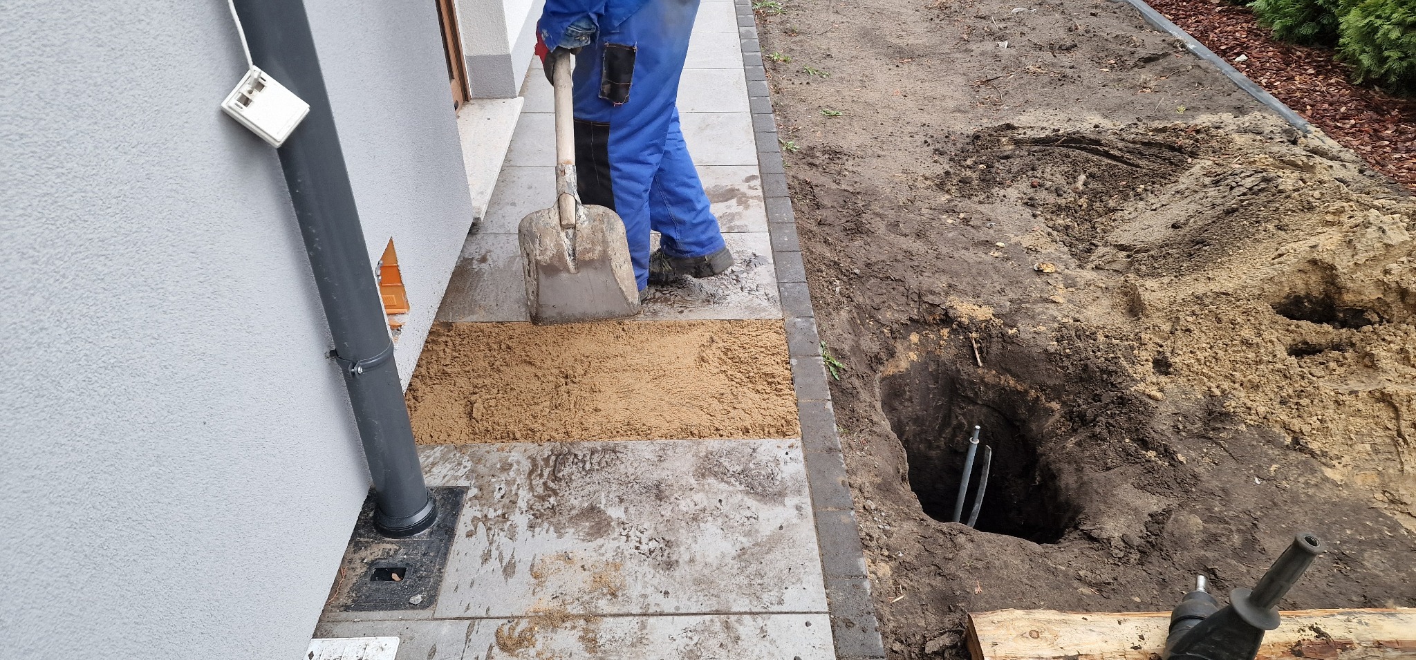 Prace ziemne przy budynku: wykop z rurami, łopata i piasek. Instalacja podziemna w trakcie realizacji, widoczne elementy instalacji elektrycznej.