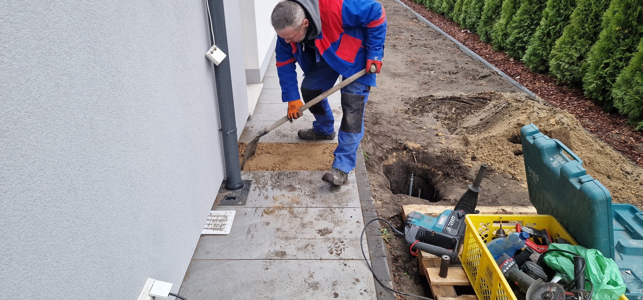Pracownik w kombinezonie kopie dół pod instalację odgromową przy budynku. Widoczne narzędzia, skrzynka z akcesoriami i fragment żywopłotu.