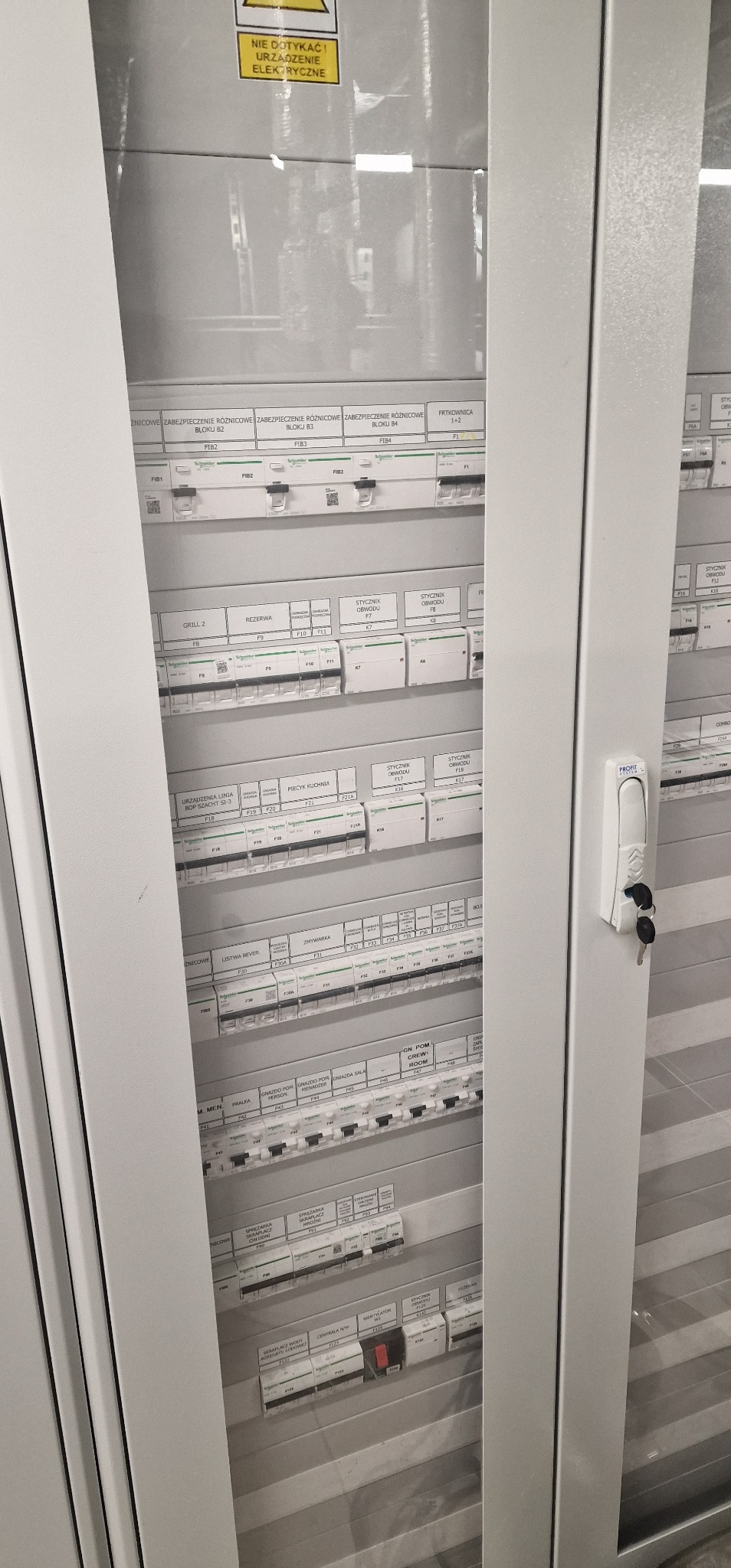 Szara szafa elektryczna z widocznymi bezpiecznikami i opisami obwodów. Kluczyki w zamku. Żółta naklejka ostrzegawcza na górze. Widok z bliska na rozdzielnicę.