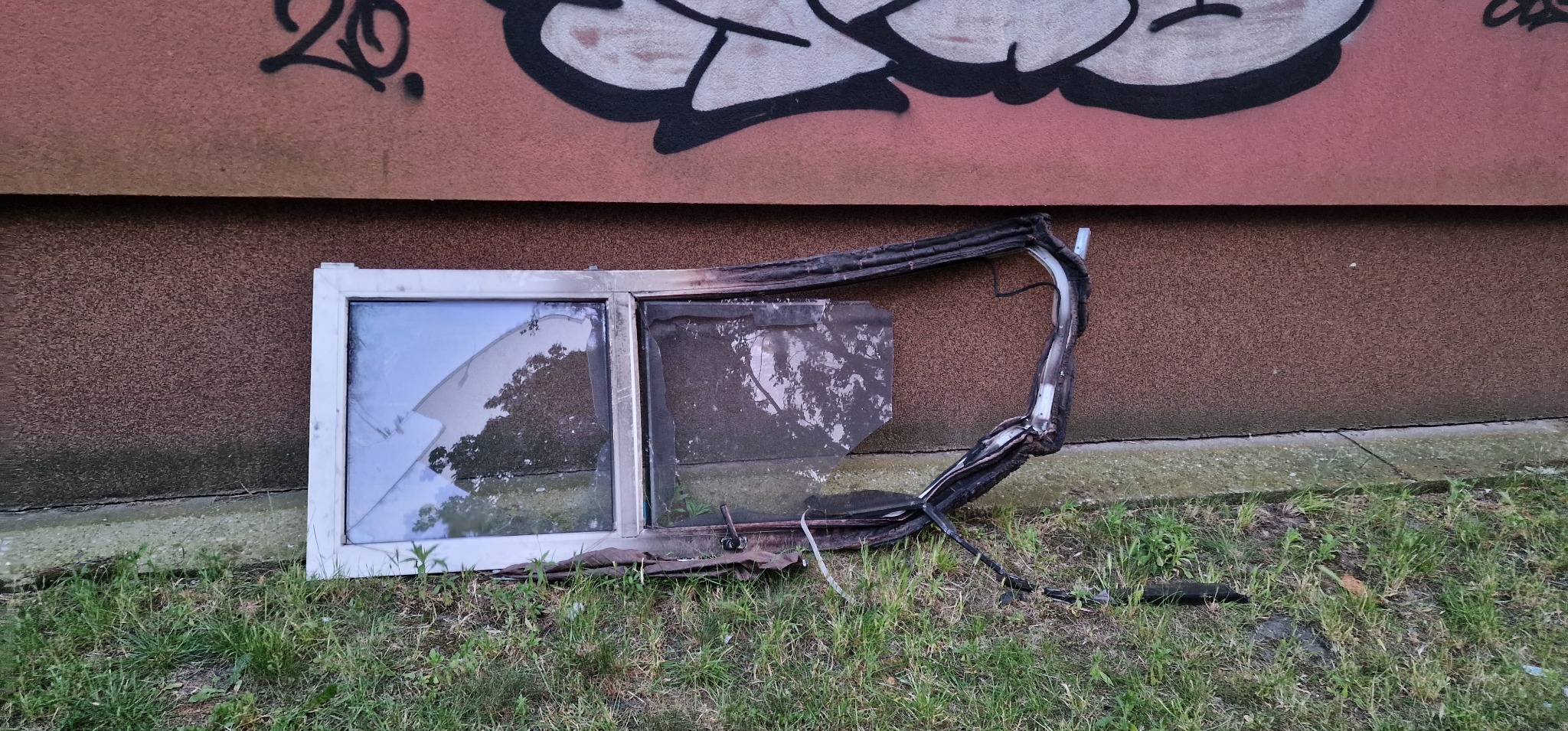 Spalone okno z uszkodzonymi szybami i ramą leży na trawie przy ścianie budynku z graffiti. Widoczne ślady pożaru i zniszczenia.