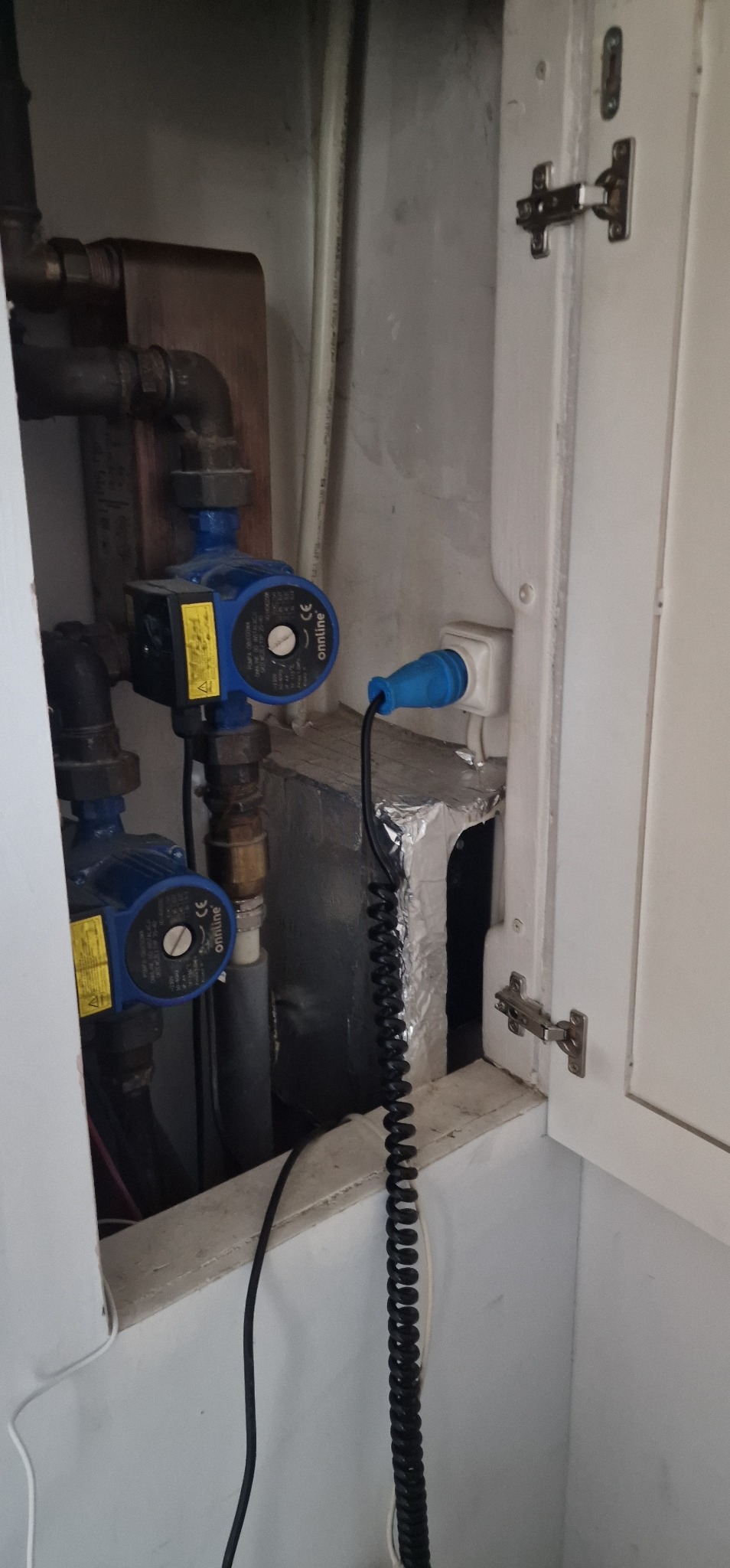 Dwie niebieskie pompy obiegowe z podłączonym przewodem zasilającym do gniazdka. Instalacja w szafce z rurami i izolacją termiczną.