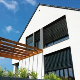 Nowoczesny dom z białymi ścianami i ciemnymi roletami zewnętrznymi, widok z dołu na tle błękitnego nieba, drewniana pergola w rogu kadru.