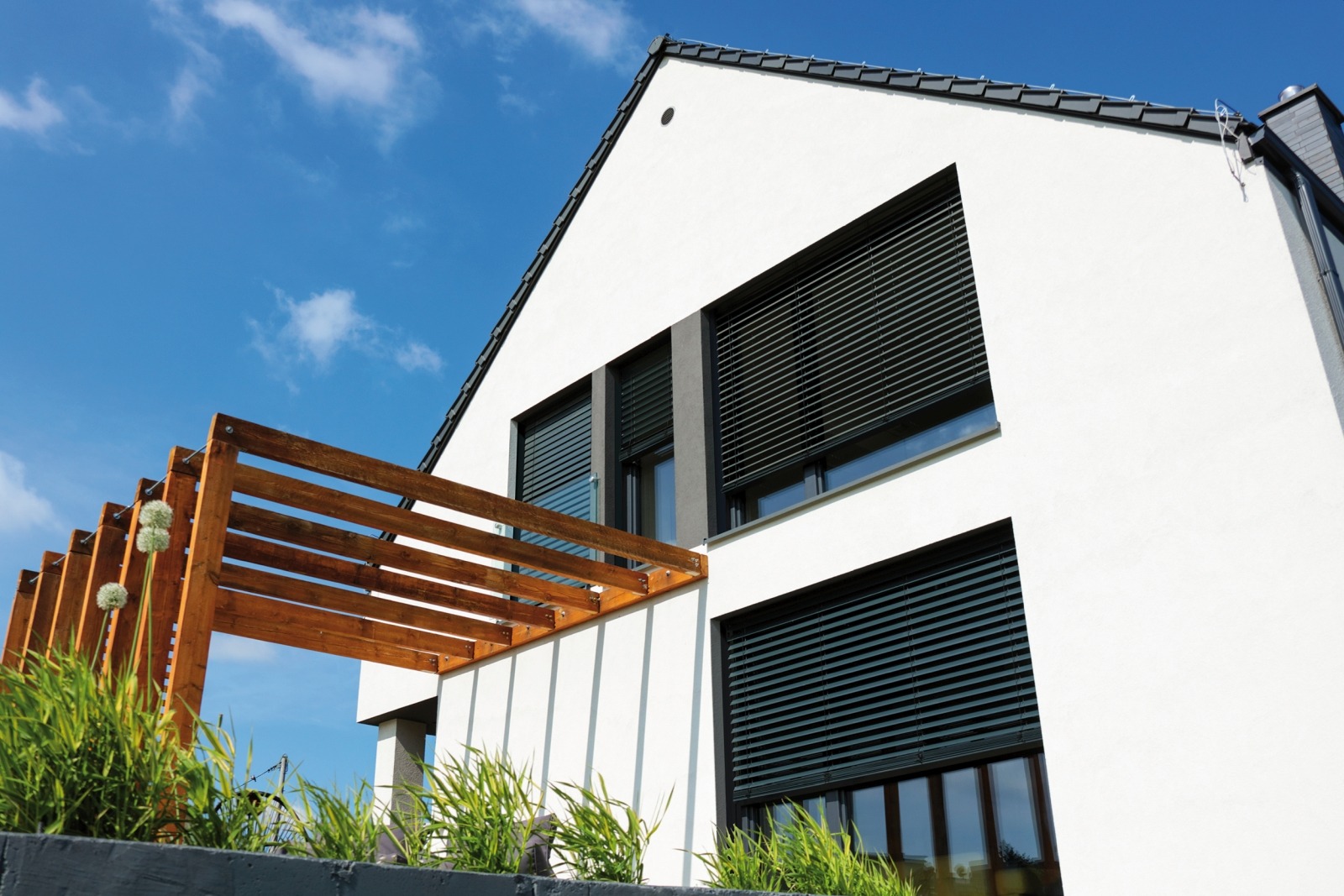 Nowoczesny dom z białymi ścianami i ciemnymi roletami zewnętrznymi, widok z dołu na tle błękitnego nieba, drewniana pergola w rogu kadru.