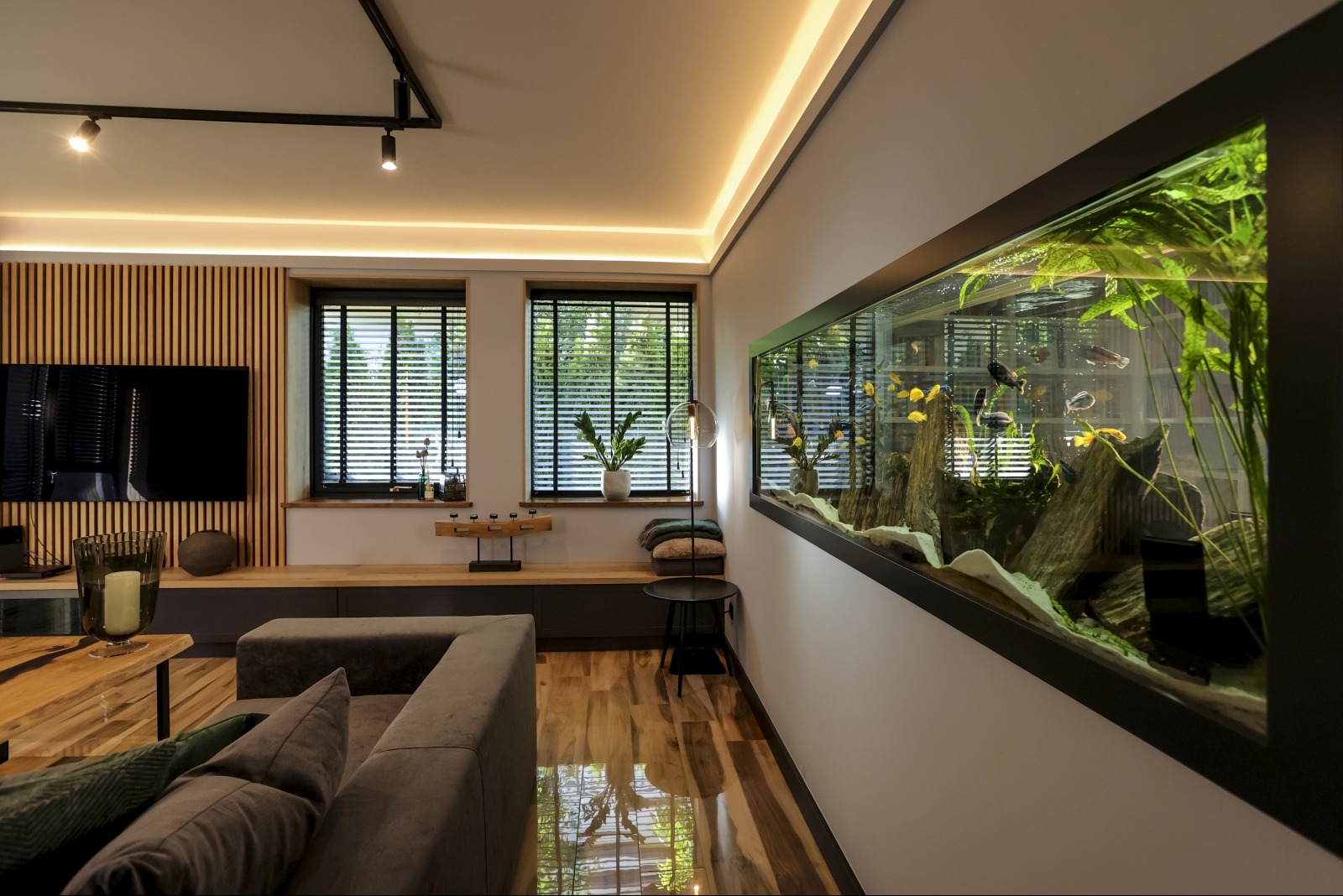 Nowoczesny salon z dużym akwarium wbudowanym w ścianę, widoczne żaluzje w oknach, drewniana ściana z telewizorem, parkiet odbijający światło.