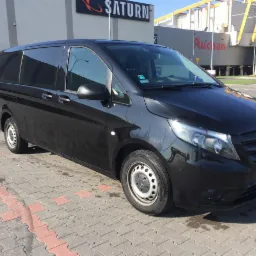 Czarny Mercedes Vito z przyciemnianymi szybami, zaparkowany na kostce brukowej przed budynkami Saturn i Auchan.