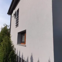 Jasnoszara elewacja domu jednorodzinnego z widocznym oknem z drewnianą ramą, satelitą na ścianie i cieniem drzew na dolnej części ściany.