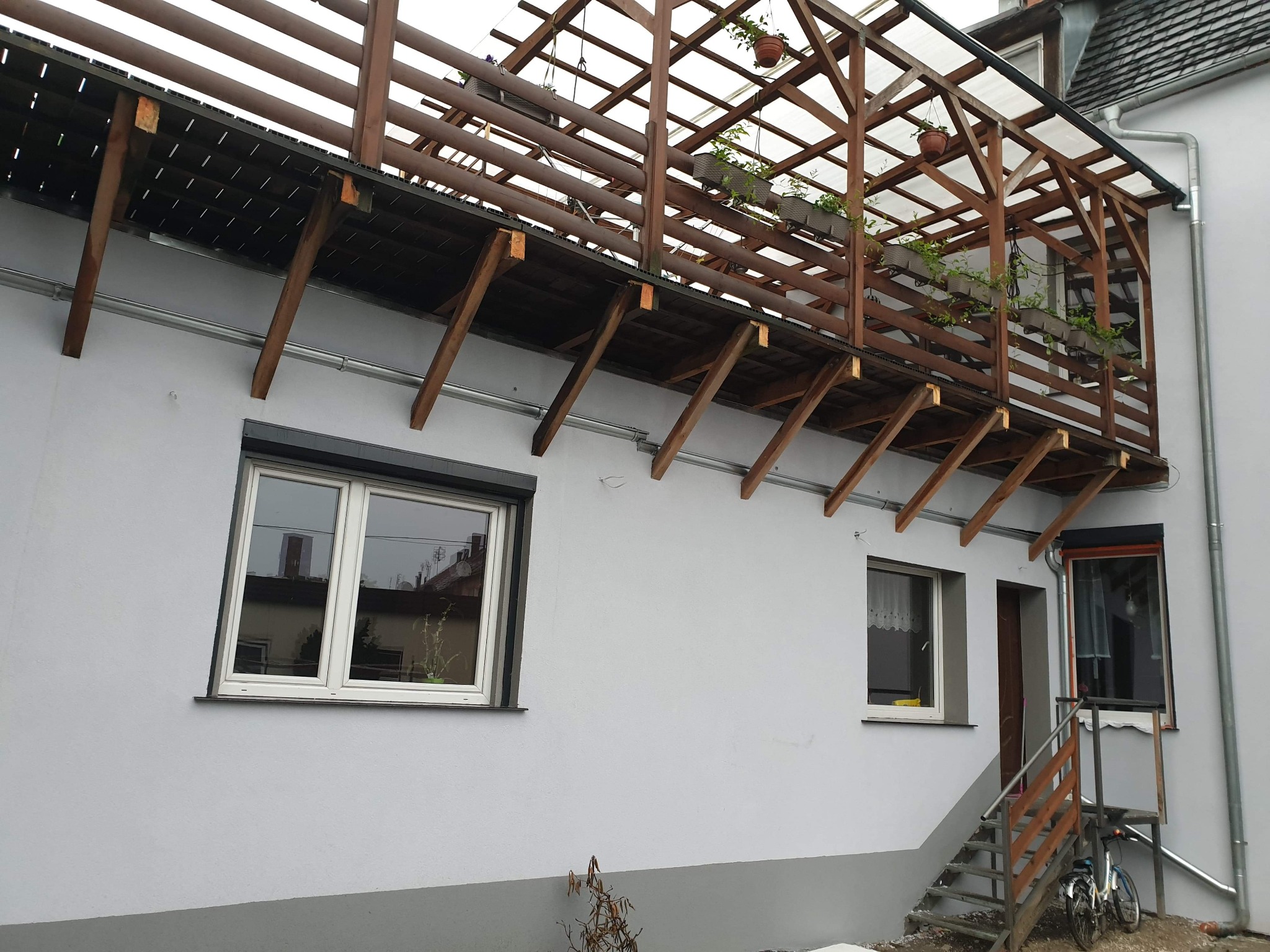 Szara elewacja budynku z drewnianym zadaszonym tarasem, widoczne okna z białymi ramami, schody wejściowe i rower.