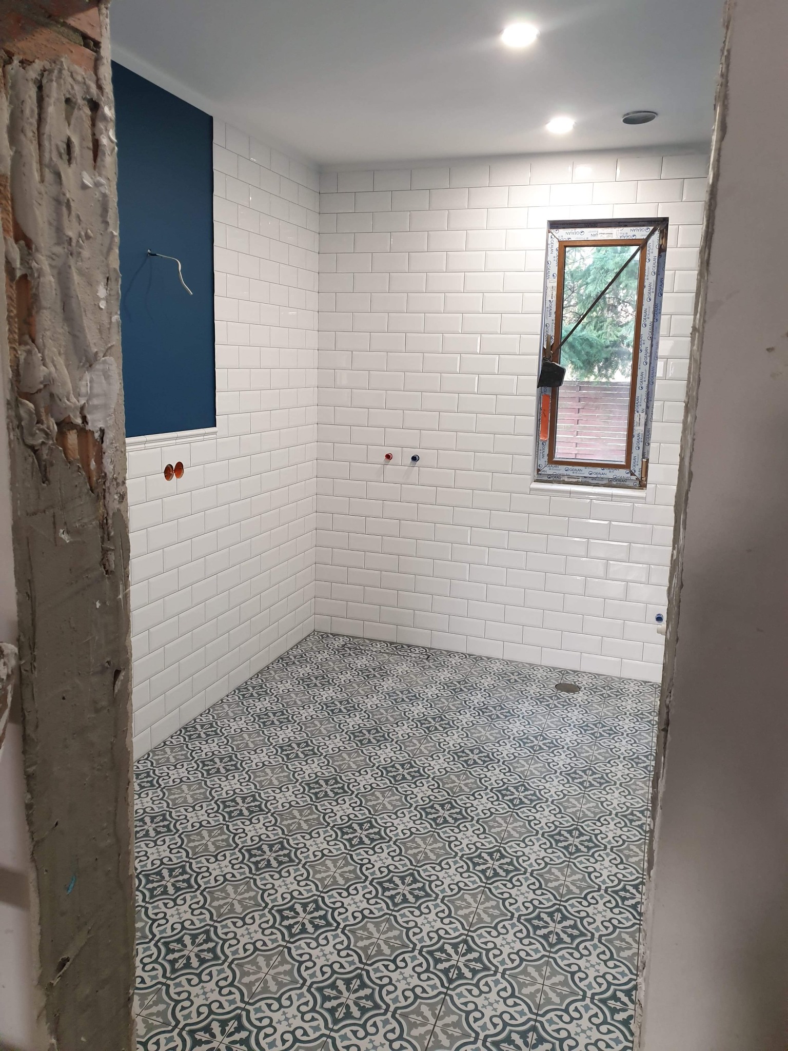 Łazienka w trakcie remontu: białe, błyszczące płytki typu metro na ścianach, wzorzysta podłoga w odcieniach szarości i błękitu, surowa ściana przy wejściu, okno z widokiem na zieleń.