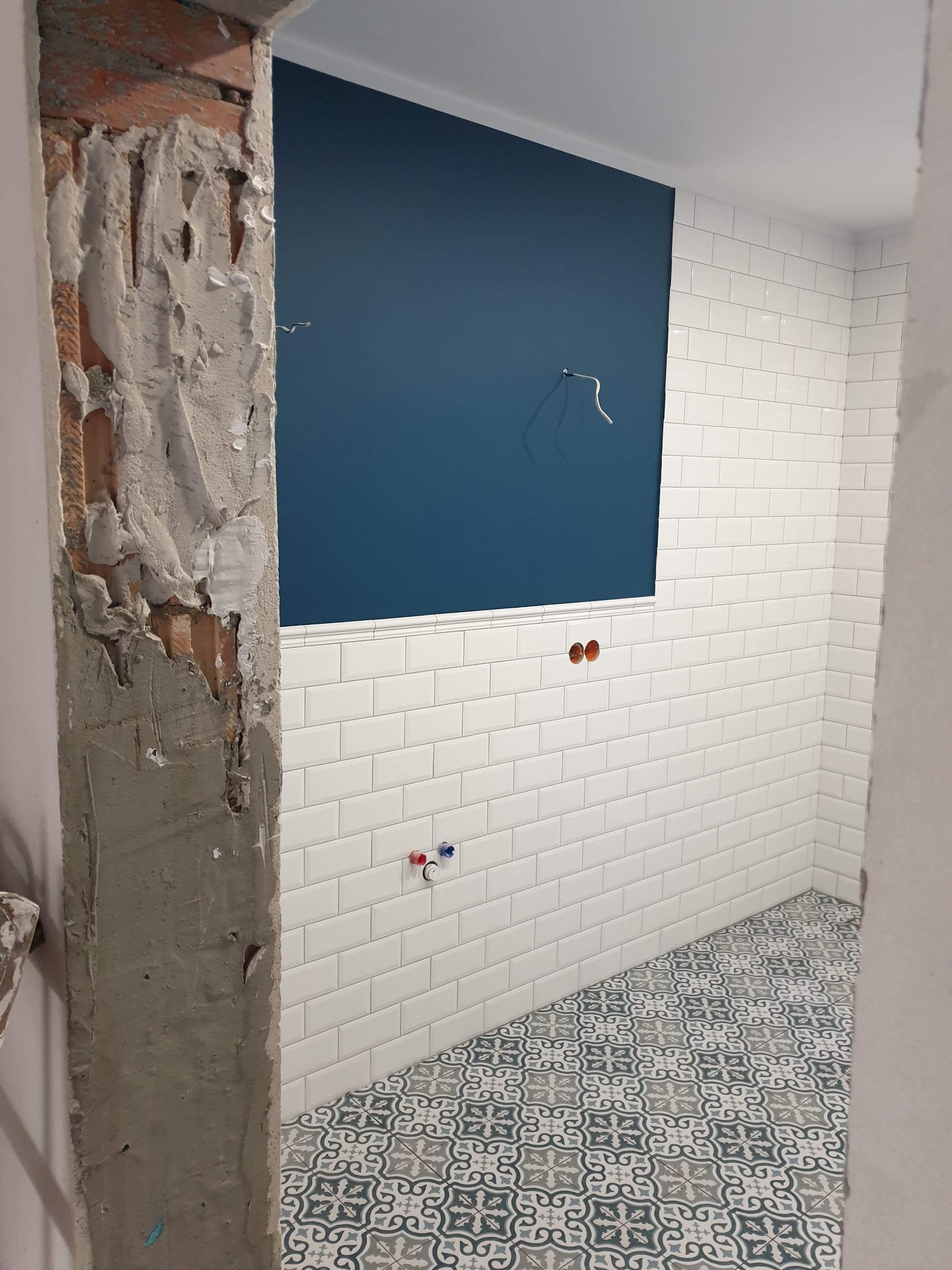 Wnętrze łazienki w trakcie prac wykończeniowych: widoczna surowa ściana z resztkami zaprawy obok ułożonych białych płytek metra na ścianie i wzorzystej podłogi, fragment niebieskiej ściany...