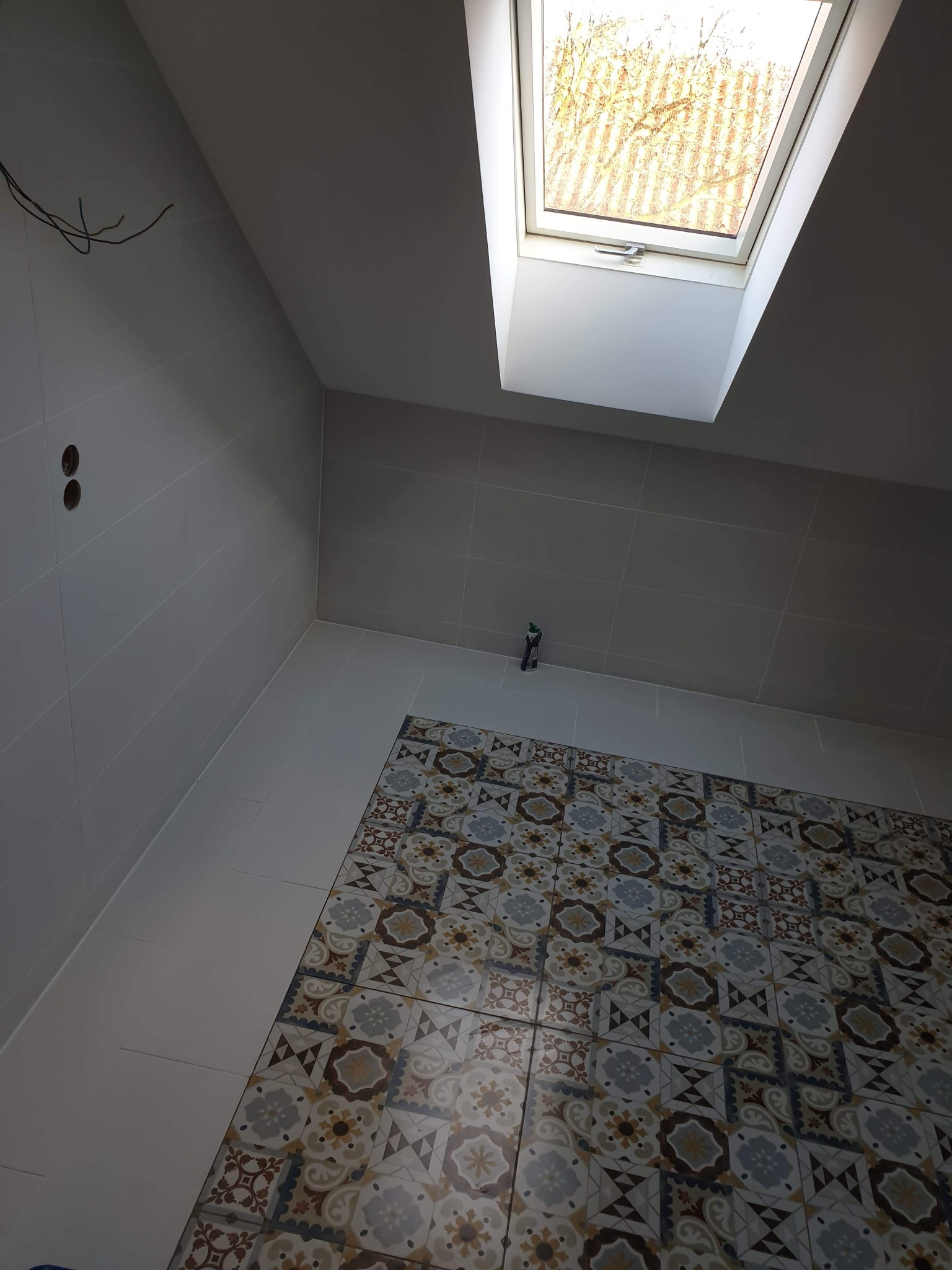 Świeżo wykafelkowana podłoga w łazience z wzorzystymi płytkami, widoczne przyłącza elektryczne i okno dachowe w skosie.