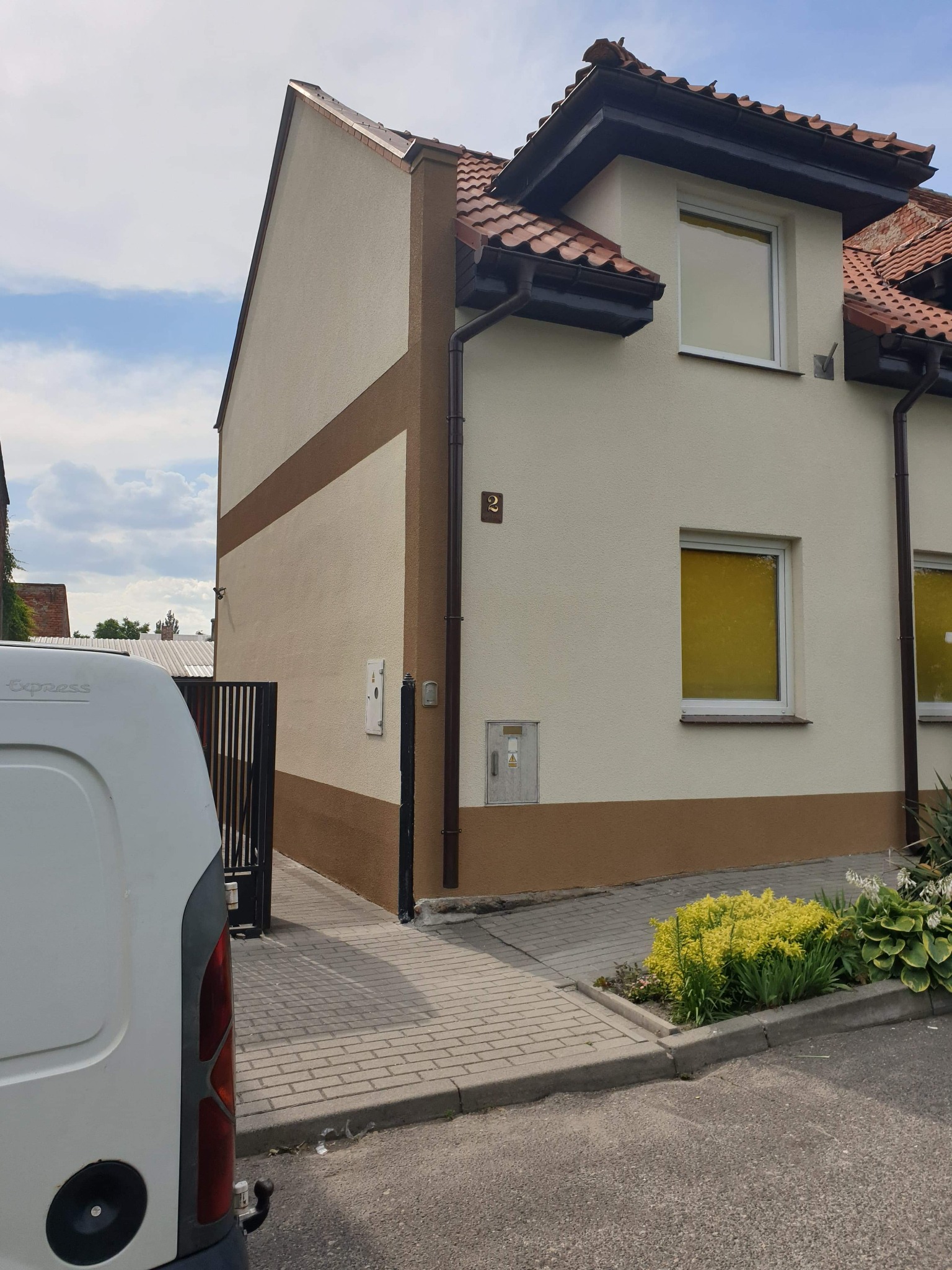 Dwukondygnacyjny dom z elewacją w kolorze beżowym i brązowym, widoczny numer domu '2', okna zakryte folią, fragment białego samochodu dostawczego na pierwszym planie.
