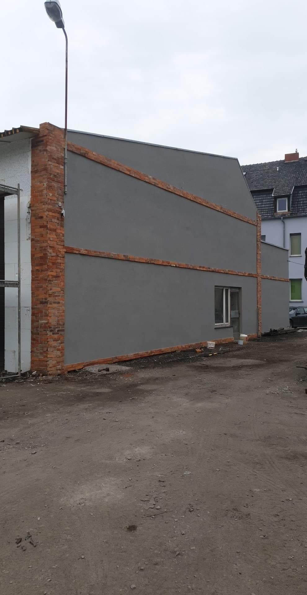 Szara elewacja budynku z ceglanymi detalami, widoczne fragmenty rusztowania i lampa uliczna, teren przed budynkiem nieutwardzony.