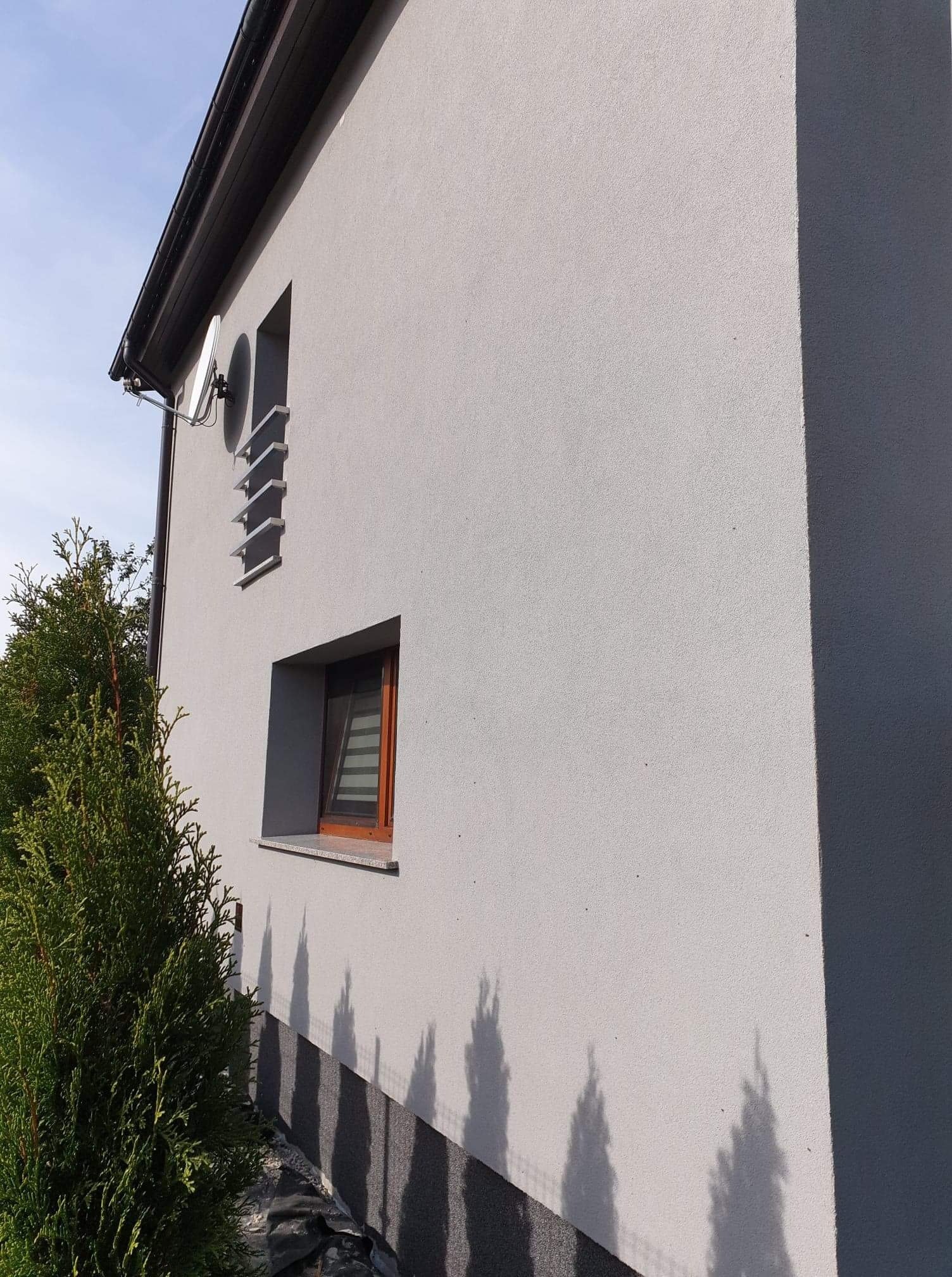 Jasnoszara elewacja domu jednorodzinnego z widocznym oknem z drewnianą ramą, satelitą na ścianie i cieniem drzew na dolnej części ściany.