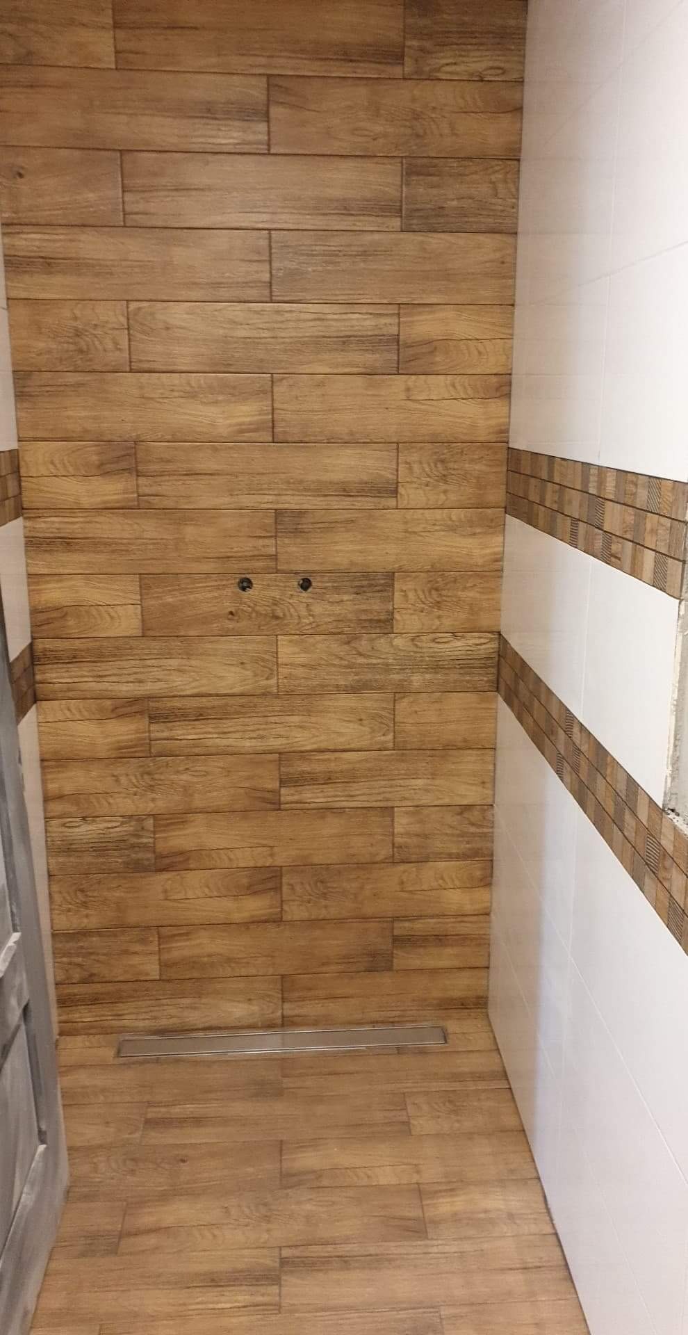 Płytki imitujące drewno na ścianie prysznica z odpływem liniowym w podłodze, widoczne przyłącza wodne, wykończenie z białych płytek z brązowym wzorem.
