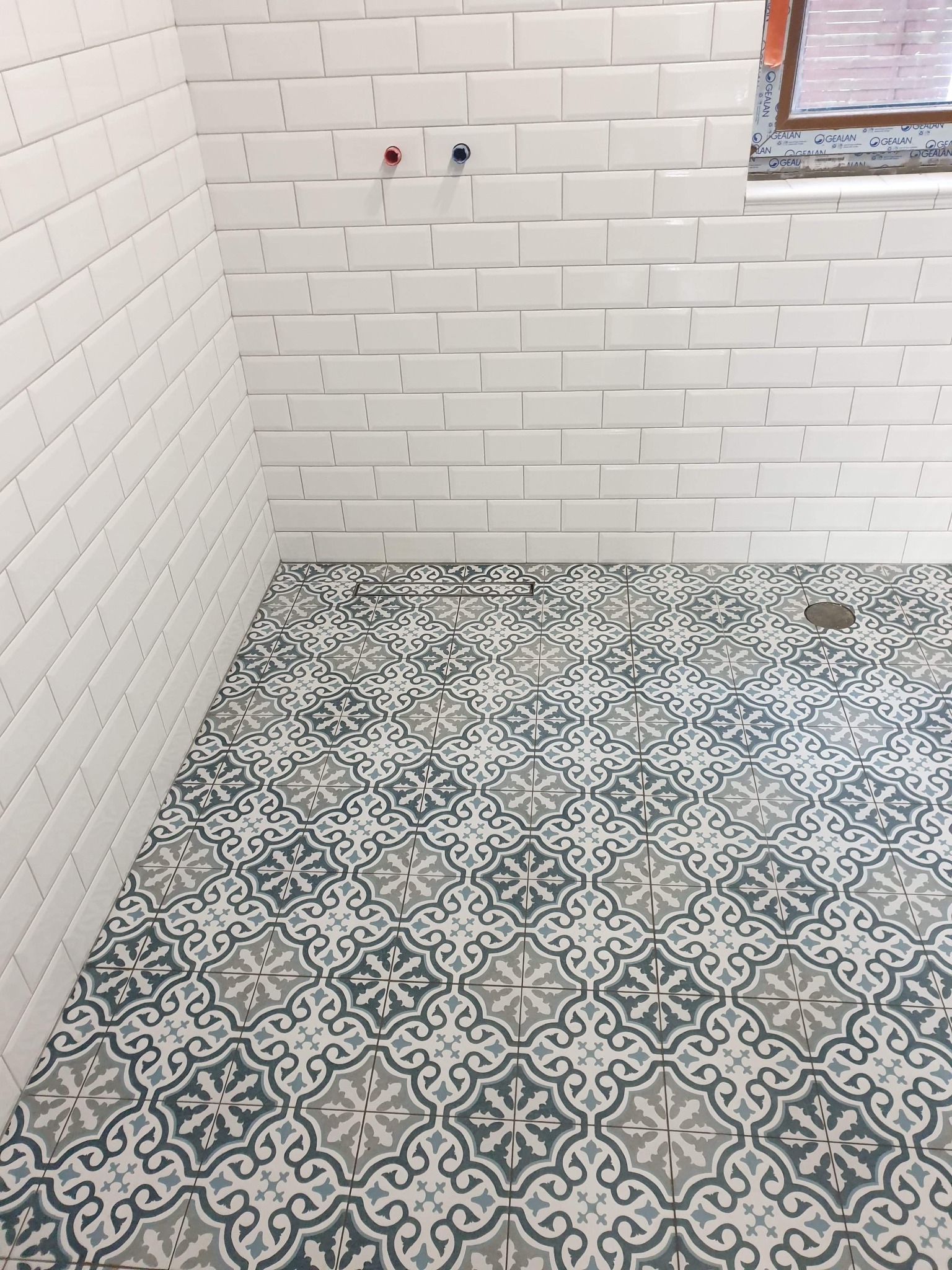 Wykończona łazienka w Krotoszynie z białymi, prostokątnymi płytkami ściennymi i wzorzystą, niebiesko-szarą podłogą z odpływem. Widoczne dwa wystające przyłącza wodne.