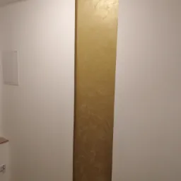 Fragment ściany z dekoracyjnym pasem w odcieniach złota i beżu, imitującym marmurkową strukturę, na tle gładkiej, białej ściany.