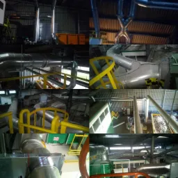 Wnętrze hali przemysłowej z systemem rur wentylacyjnych, ż&oacute;łtymi barierkami i kontenerami wypełnionymi materiałem izolacyjnym, widok z g&oacute;ry na instalację przemysłową.