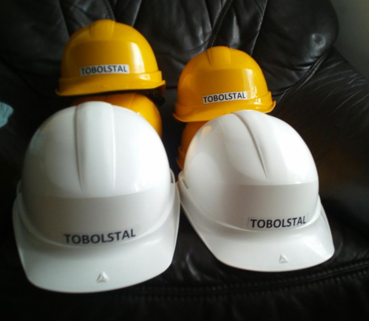 Stos żółtych i białych kasków budowlanych z logo 'TOBOLSTAL' na skórzanym tle.