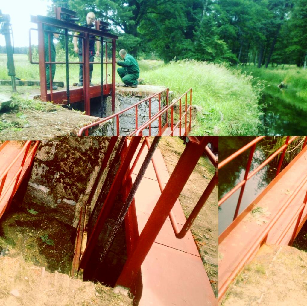 Prace konserwacyjne przy metalowej konstrukcji śluzy wodnej, trzech pracowników w kombinezonach roboczych przy czerwonej bramie śluzy na tle zieleni i wody.