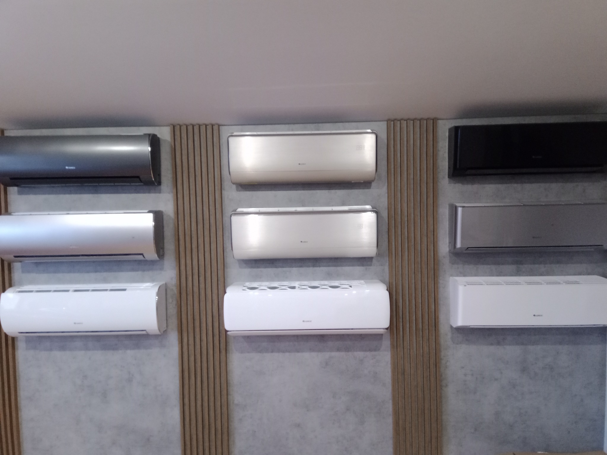 Ekspozycja różnych modeli klimatyzatorów ściennych w kolorach: szarym, srebrnym, białym i czarnym, zamontowanych na ścianie z dekoracyjnymi panelami pionowymi.