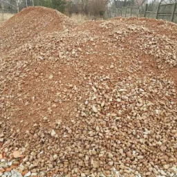 Dwie pryzmy drobnego, brązowo-czerwonego kruszywa na placu budowy, z fragmentami gruzu i cegieł w dolnej części kadru, widoczne drzewa w tle.