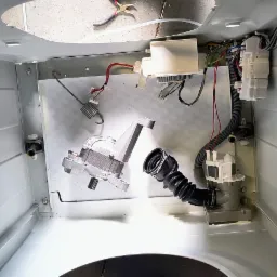 Wnętrze pralki automatycznej w trakcie naprawy, widoczne elementy: silnik, pompa odpływowa z gumowym wężem karbowanym, moduł sterujący z przewodami, sprężyna, kombinerki leżące na górnej krawędzi...