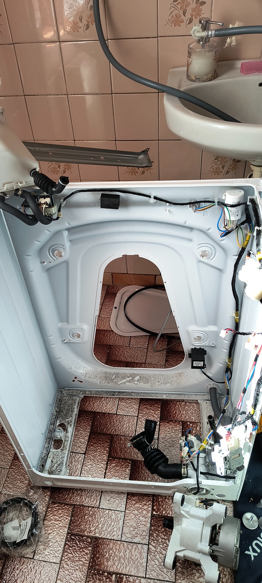 Wnętrze pralki automatycznej podczas naprawy, widoczne okablowanie, pompa i inne elementy, tło stanowią brązowe kafelki łazienkowe.