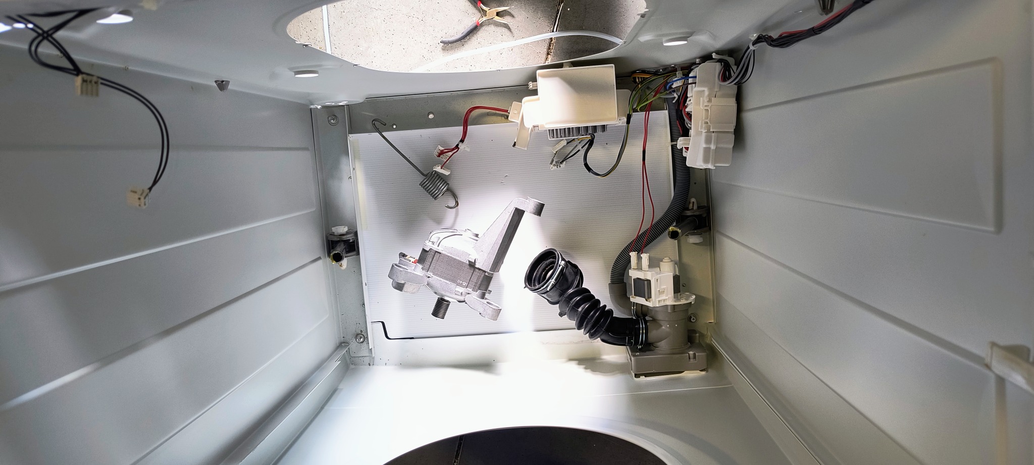 Wnętrze pralki automatycznej w trakcie naprawy, widoczne elementy: silnik, pompa odpływowa z gumowym wężem karbowanym, moduł sterujący z przewodami, sprężyna, kombinerki leżące na górnej krawędzi...