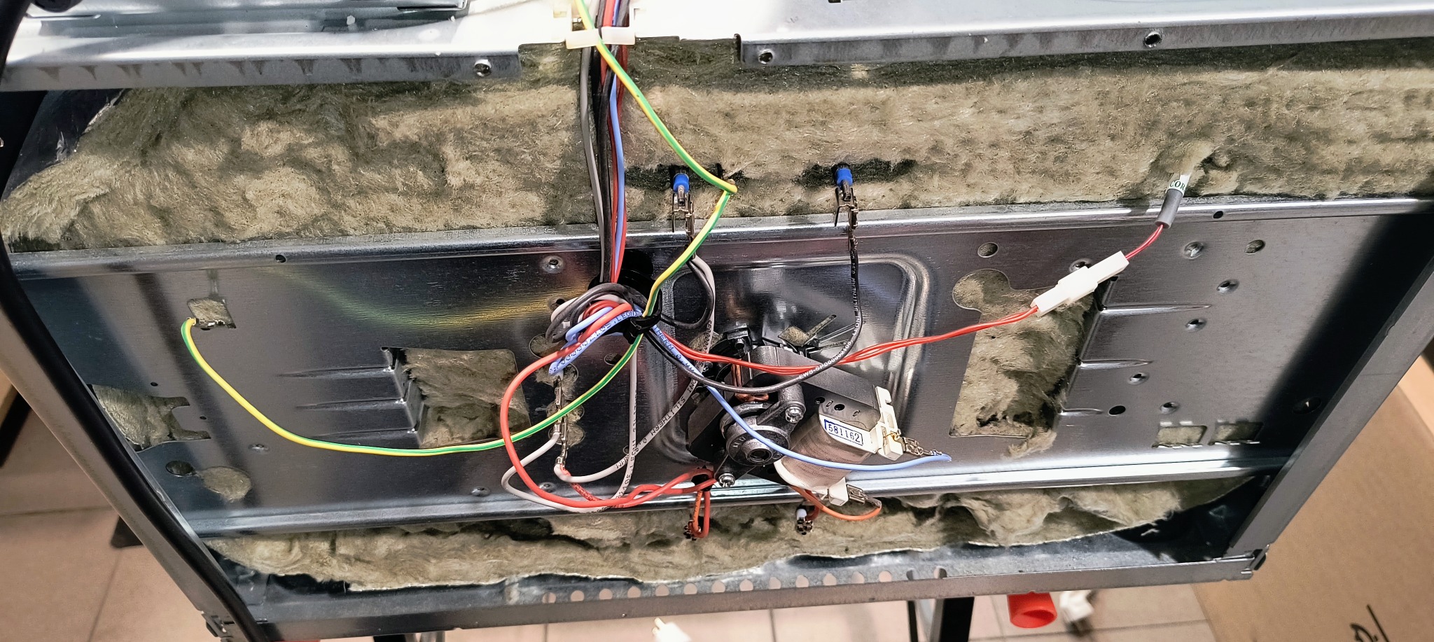 Wnętrze piekarnika z widoczną izolacją, metalową obudową oraz wiązką kabli i elementami elektrycznymi podczas naprawy.