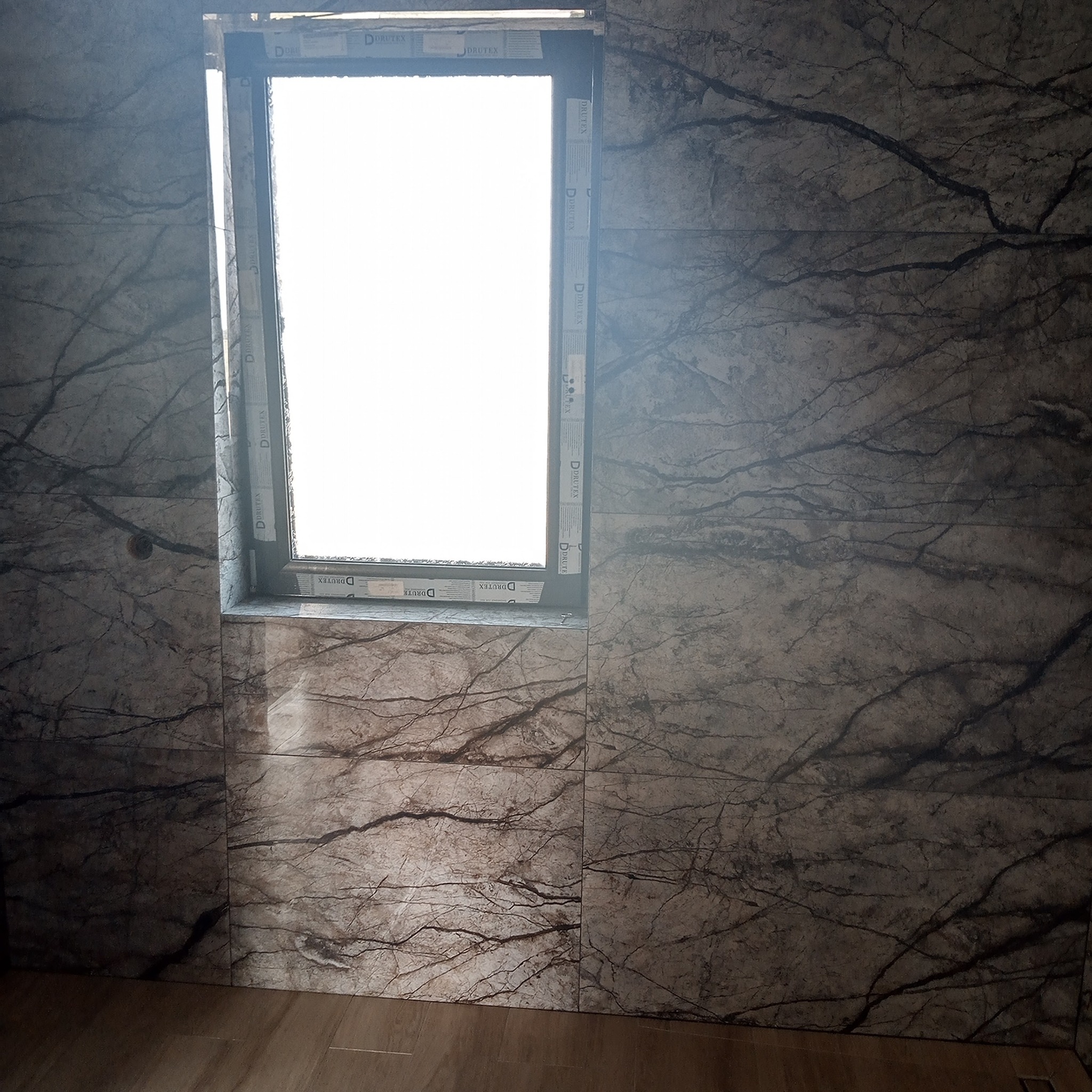 Ściana wyłożona płytkami imitującymi ciemny marmur z wyraźnym wzorem żyłkowania, z oknem w centralnej części kadru, z widocznymi elementami ramy okiennej z nadrukowanym logo producenta.
