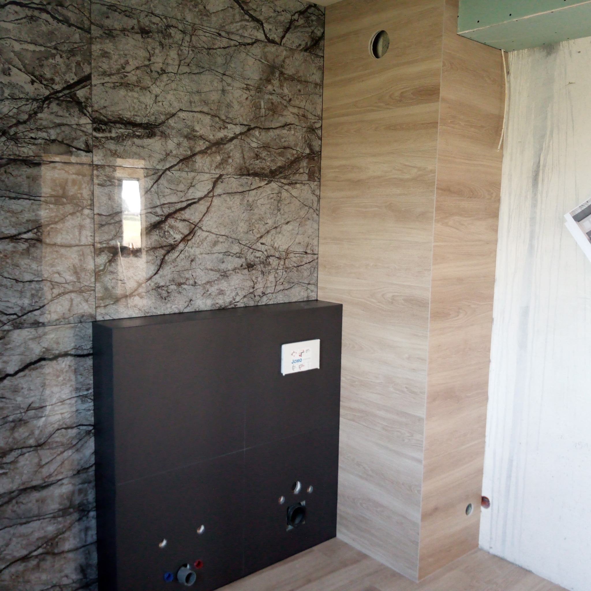 Wykończona ściana w łazience z marmurowymi i drewnopodobnymi płytkami, widoczna zabudowa stelaża WC z wyprowadzonymi przyłączami wodnymi i kanalizacyjnymi, surowy stan ściany po prawej stronie.