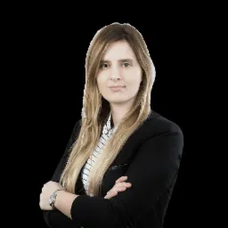 Portret kobiety w czarnej marynarce i koszuli w paski, z założonymi rękami i zegarkiem na nadgarstku, na czarnym tle.