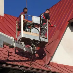 Dwóch robotników na podnośniku maluje czerwoną blachodachówkę na dachu budynku w słoneczny dzień. Jeden z nich używa pistoletu natryskowego.