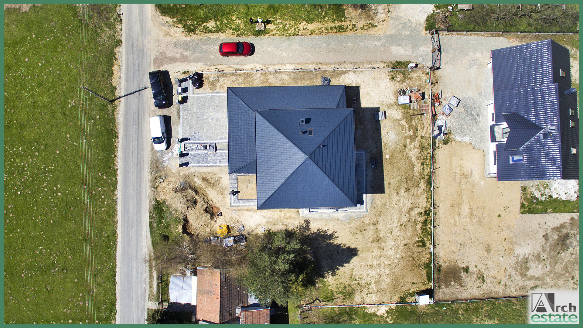 Widok z lotu ptaka na nowo wybudowany dom z ciemnoszarym dachem, częściowo wyłożonym podjazdem z kostki brukowej, placem budowy w trakcie prac wykończeniowych i dwoma zaparkowanymi samochodami...