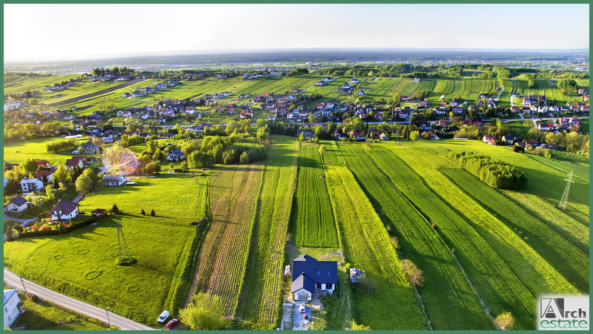 Panoramiczny widok z drona na wiejski krajobraz z nowym domem jednorodzinnym z ciemnym dachem, otoczonym zielonymi polami i zabudowaniami mieszkalnymi w oddali.