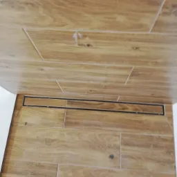 Ułożone płytki podłogowe imitujące drewno w łazience, z liniowym odpływem prysznicowym. Widoczne fugi i detale struktury płytek. Na jednej z płytek widoczny ślad po ołówku.