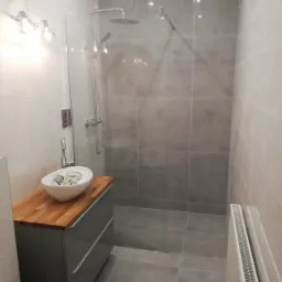 Nowoczesna łazienka z szarymi płytkami, kabiną prysznicową typu walk-in, umywalką nablatową na drewnianym blacie i grzejnikiem żeberkowym.