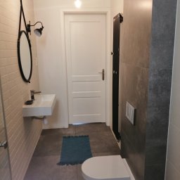 Home-Studio.pl - Nowoczesna łazienka z wiszącą toaletą, umywalką nablatową, okrągłym lustrem w czarnej ramie i grzejnikiem drabinkowym w ciemnym kolorze, oświetlona żarówkami Edisona.