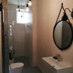 Home-Studio.pl - Nowoczesna łazienka z białymi cegiełkowymi płytkami, wiszącą toaletą, szklaną kabiną prysznicową, okrągłym lustrem w czarnej ramie i wiszącą umywalką z minimalistyczną baterią.