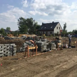 Budowa fundamentów domu jednorodzinnego: pracownik układa bloczki betonowe na przygotowanym terenie, widoczne drewniane szalunki i narzędzia, w tle dom w trakcie budowy i zaparkowany niebieski...