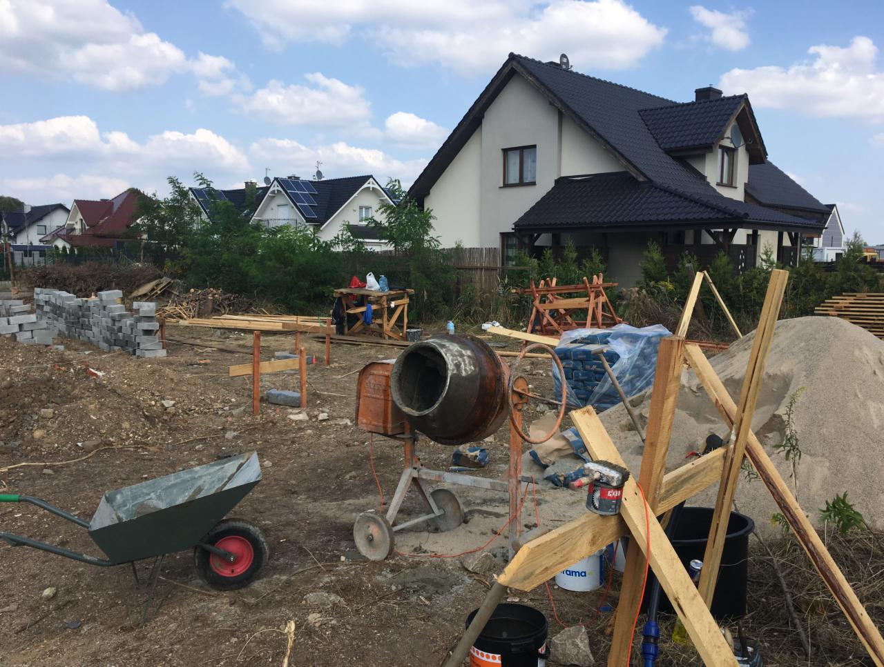 Budowa domu jednorodzinnego w trakcie realizacji: widoczny teren budowy z mieszarką do betonu, stertą bloczków betonowych, piasku, workami z cementem oraz częściowo ukończony dach na tle zabudowy...